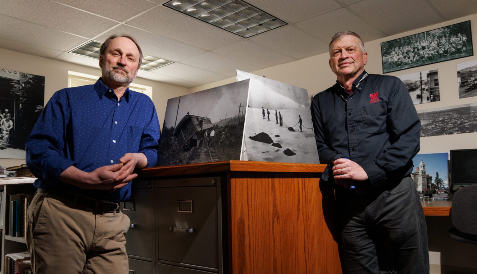 Todd Warger and Archivist Jeff Jewell leaning against the wooden desk showcasing propped up vintage photos of Whatcom.