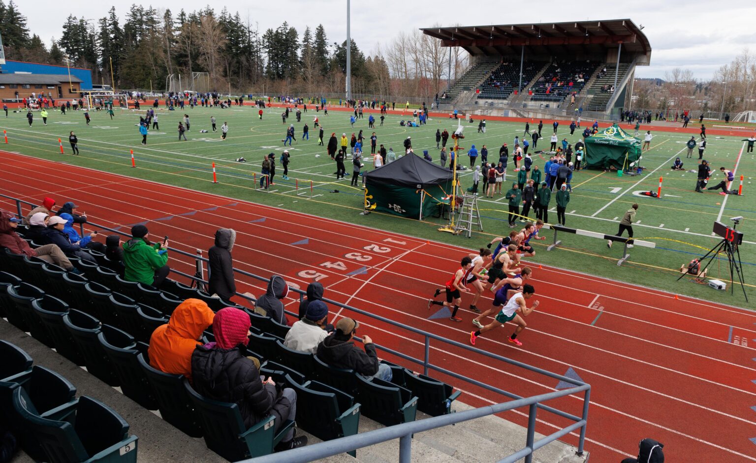 Runners take off in the boys 800-meter run as spectators on the bleacher seats watch while other spectators on the grass walk around in preparation.