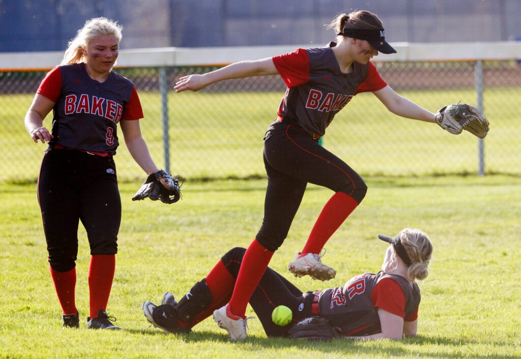 Mount Baker catcher, Lauren Valum, leaps over teammate who is on the grassy ground next to the ball as another teammate watches from the side.
