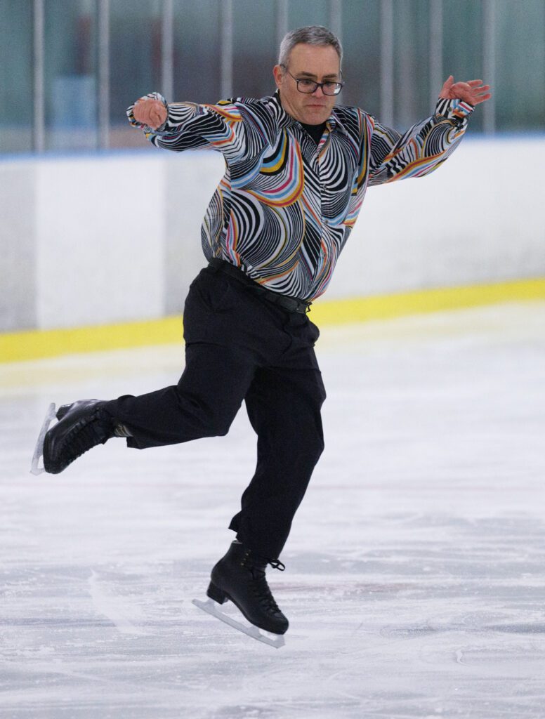 Special Olympian Scott Orvik leaps off and onto one foot as he practices on the ice rink while wearing a multicolored shirt.
