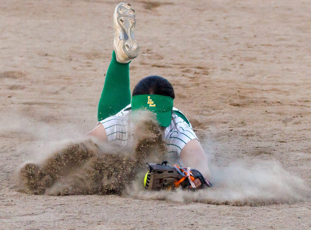 Lynden’s Campbell DeJong dives for the ball as dirt flies up into their face and all around their body as they slide across the dirt floor.