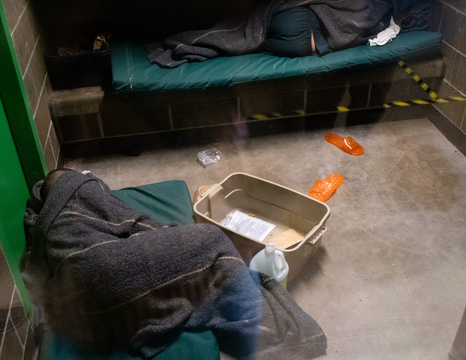 Two people under blankets sleep on green pads in a cemented jail sell. A plastic tub and orange slippers are sprawled across the cement floor.