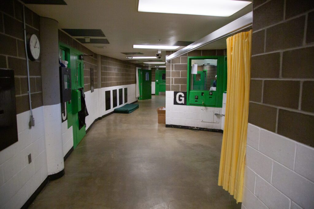 A jail hallway includes a cement floor, white and brown walls, and green doors and windows.