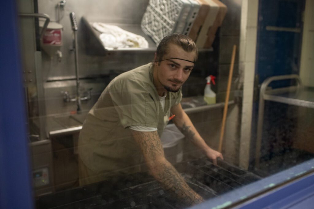 An inmate wearing a khaki uniform and hair net does dishes in a jail kitchen.