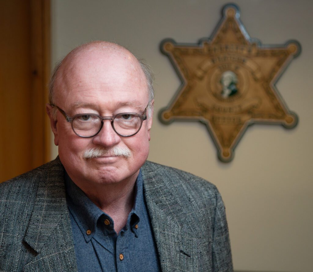 A man with a mustache and glasses poses for a photo with a sheriff star in the background.