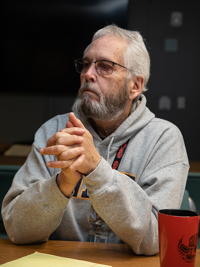 A man with a beard wearing a grey sweatshirt and glasses crosses his hands at a desk.