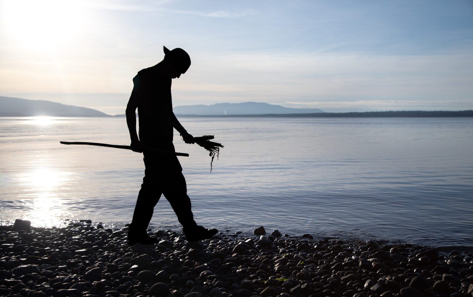 A silhouetted figure walks along a rocky beach as the sun shines over the water behind him.
