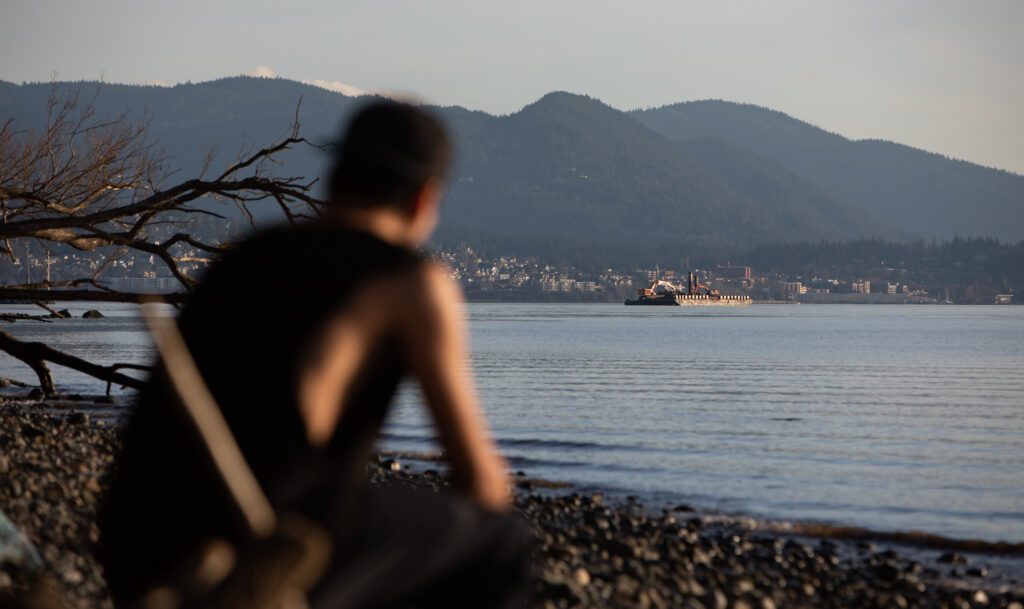 A silhouetted man sits on a rocky beach and looks out to the water and city skyline.