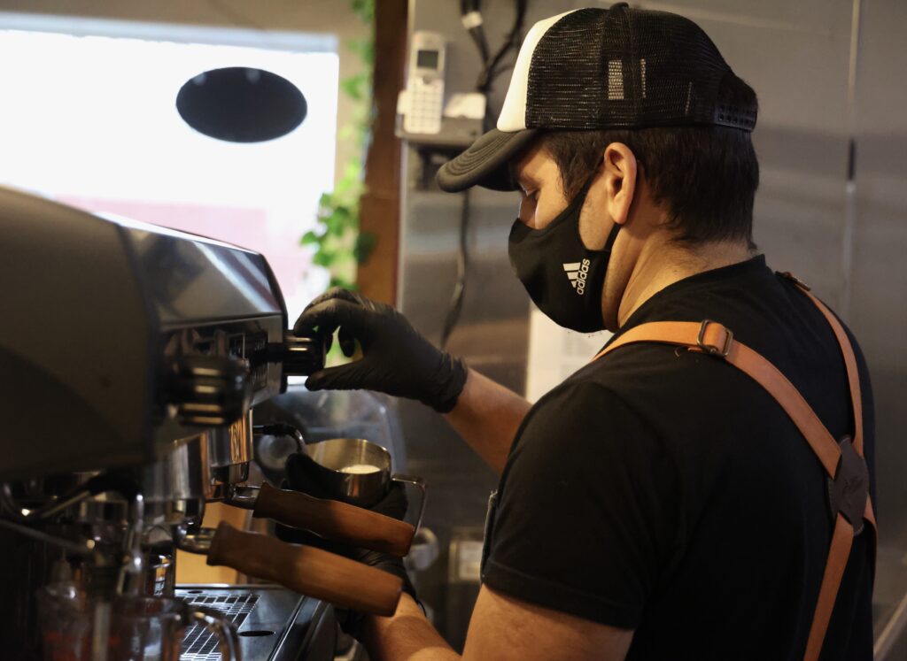 A man in a mask and hat steams milk at an espresso machine.