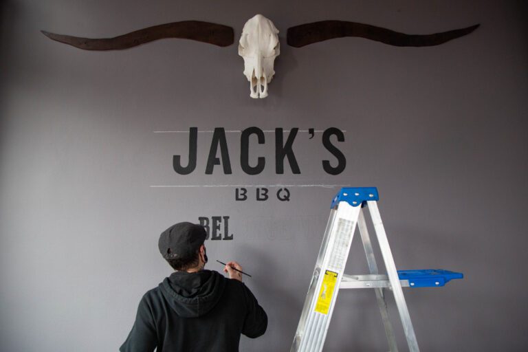 Underneath a an animal skull and a freshly painted "Jack's BBQ" a man begins to paint "Bellingham" on a grey wall.
