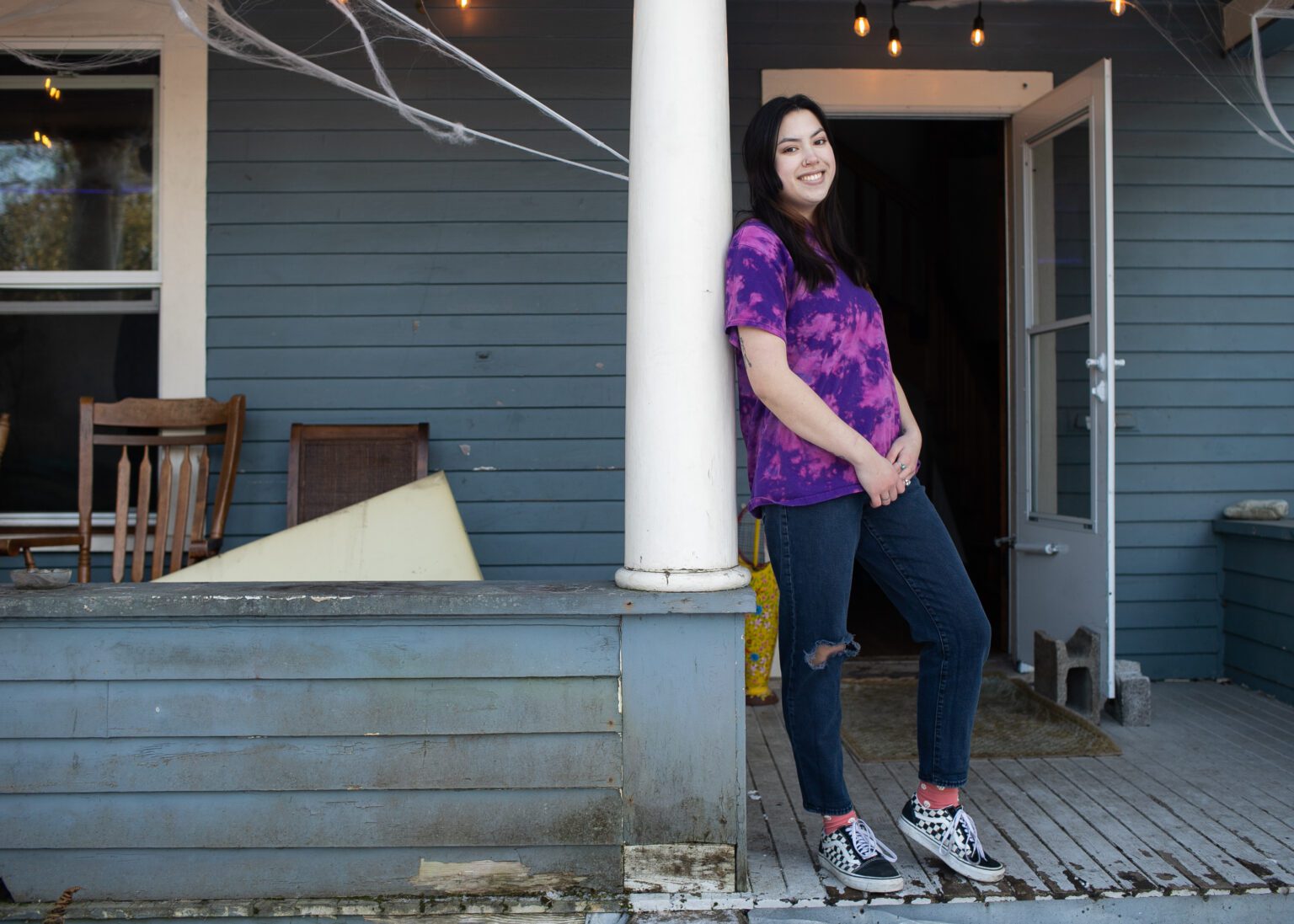 A young woman wearing a purple and pink tie dye shirt stands on the porch of a dingy blue house.