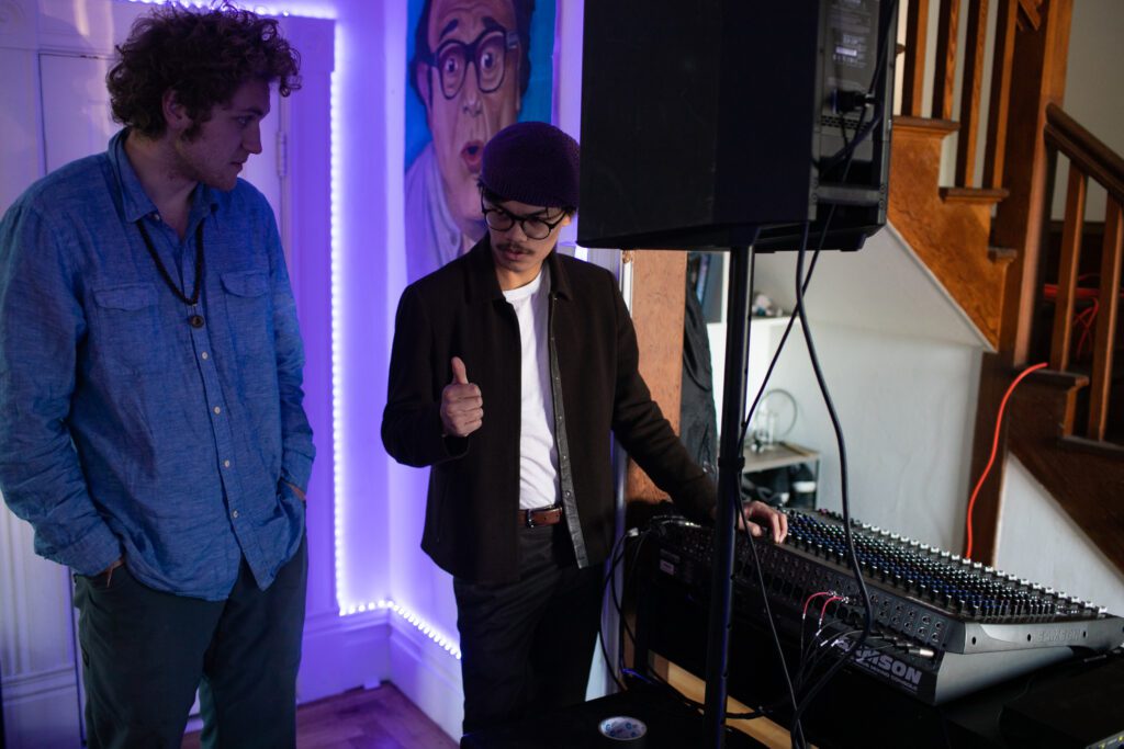 Two men stand beside a speaker and a mixing board.