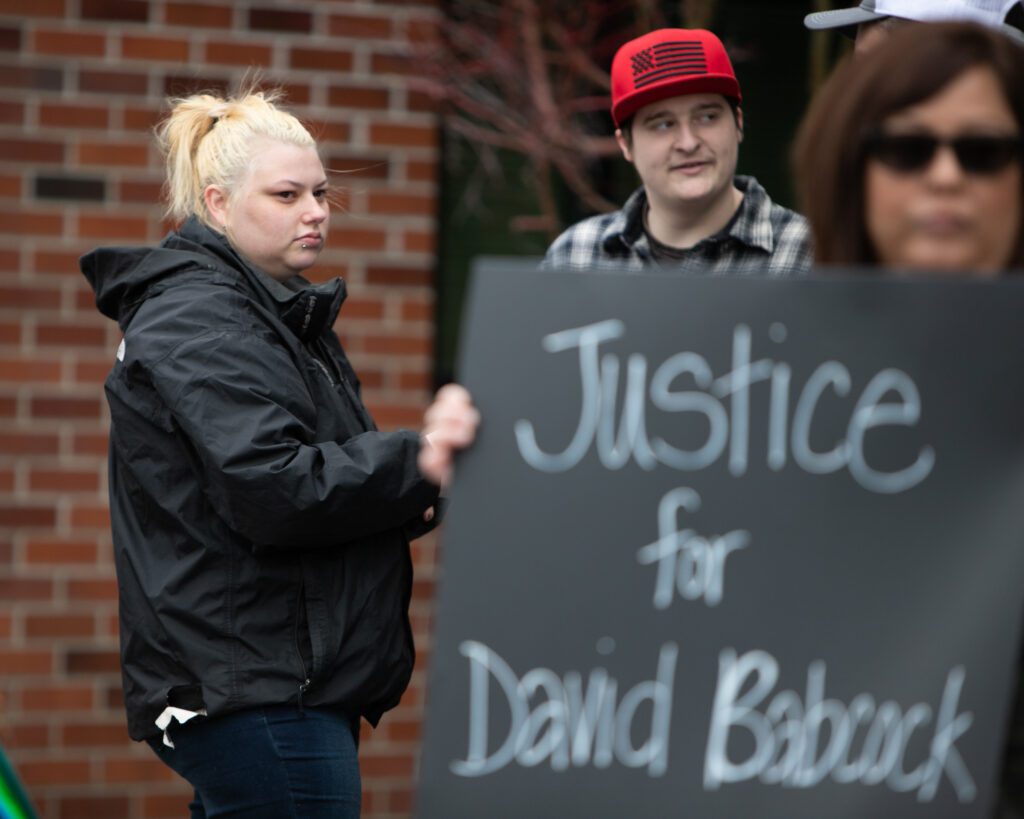 Elizabeth Babcock attending a protest outside of Sedro-Wooley Police Department, behind a woman holding up a sign called Justice for David Babock.