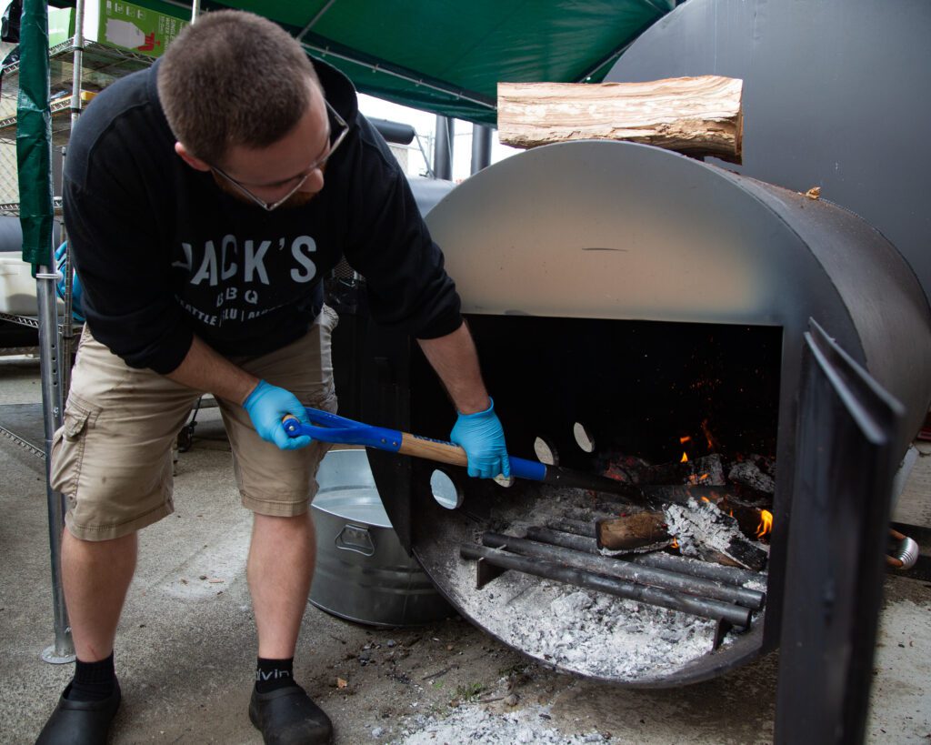 A man in a Jack's BBQ sweatshirt pokes logs and flames in a smoker outside.
