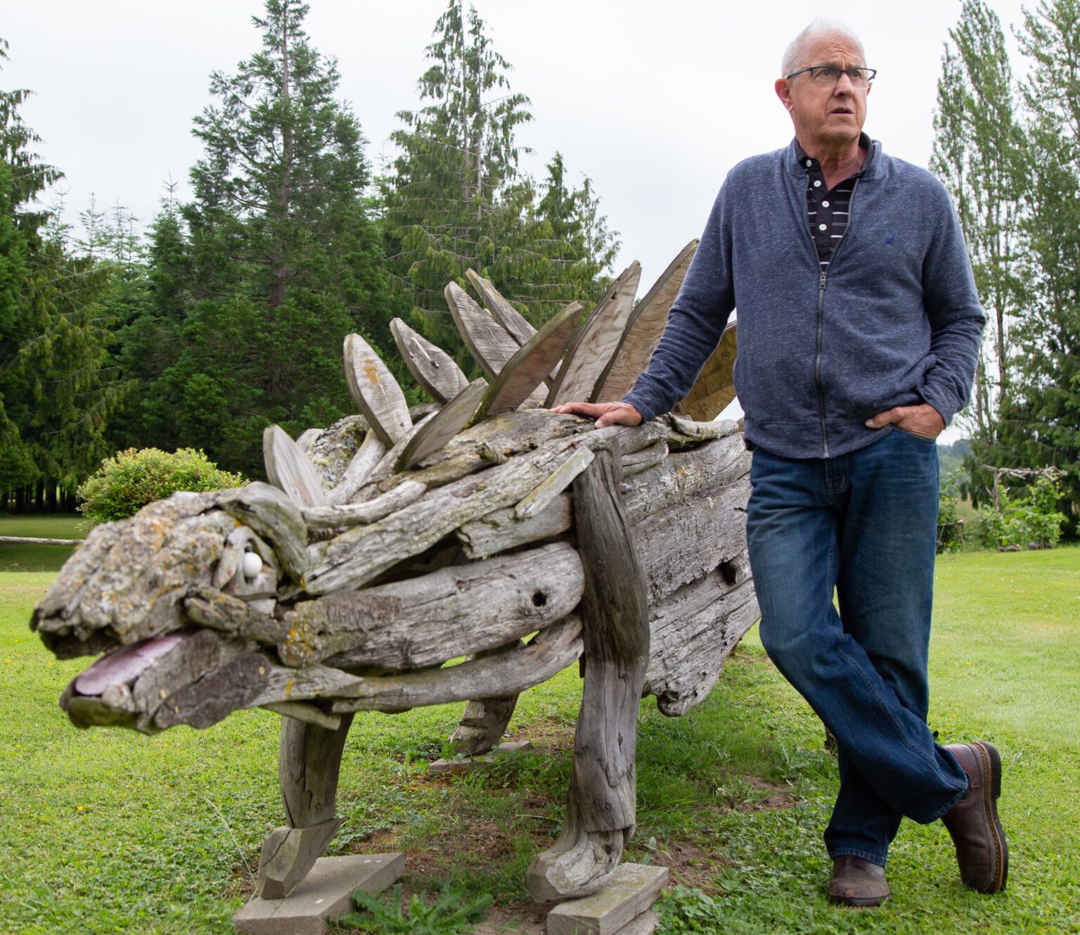 Joe Treat leans against his driftwood stegosaurus sculpture on display outside of his home in Bow on July 1. Other animals at Bowrassic Park include a horse