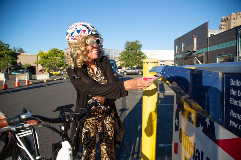 Patti Braimes puts in her ballot in a drop box as she smiles while holding onto her bicycle and wearing a multicolored helmet.