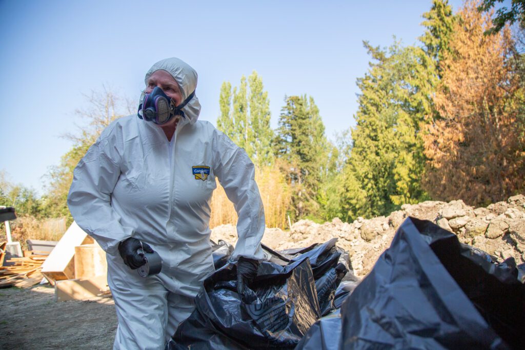 A woman wearing a gas mask and protective gear tape a garbage bag shut.