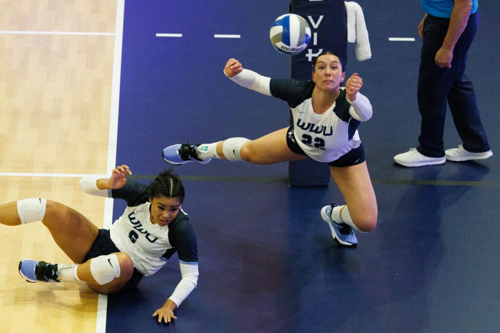 A volleyball team member dives around their teammate that's aiming to hit the ball.