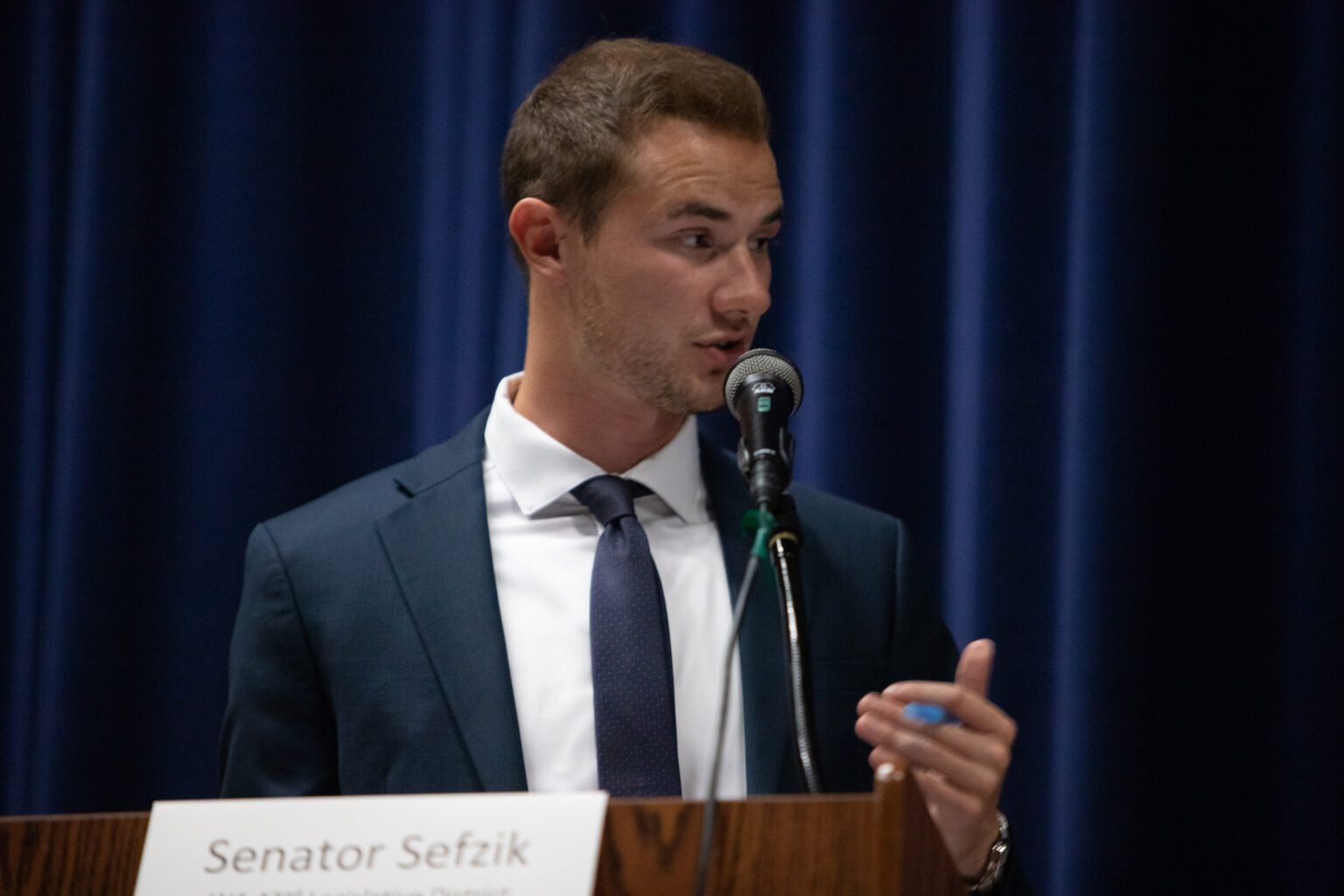 Sen. Simon Sefzik speaks through a microphone with a dark blue backdrop behind him.