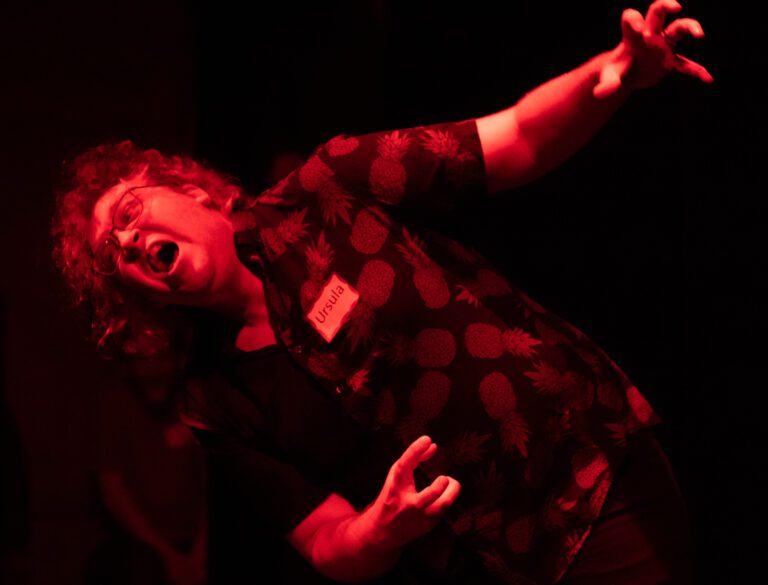 Michael Nelson-Brannan, as Ursula the spa owner pretends to die on stage while wearing a pineapple print shirt.