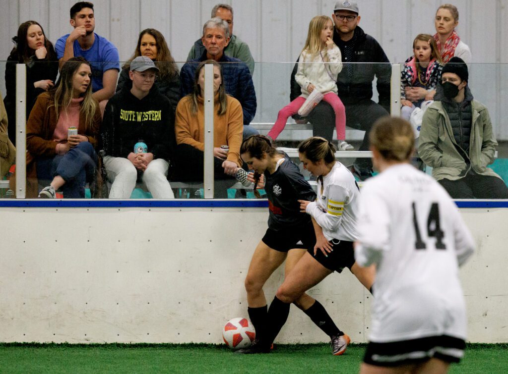 Two women battle for the soccer ball, one of them slightly leaning against the protective barrier in front of the audience on the bleachers.