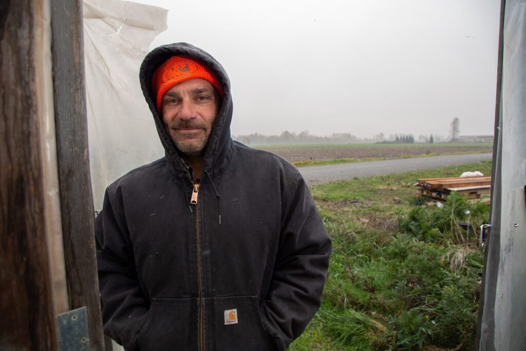 A farmer standing in a doorway of a greenhouse wearing a hoodie and an orange beanie.