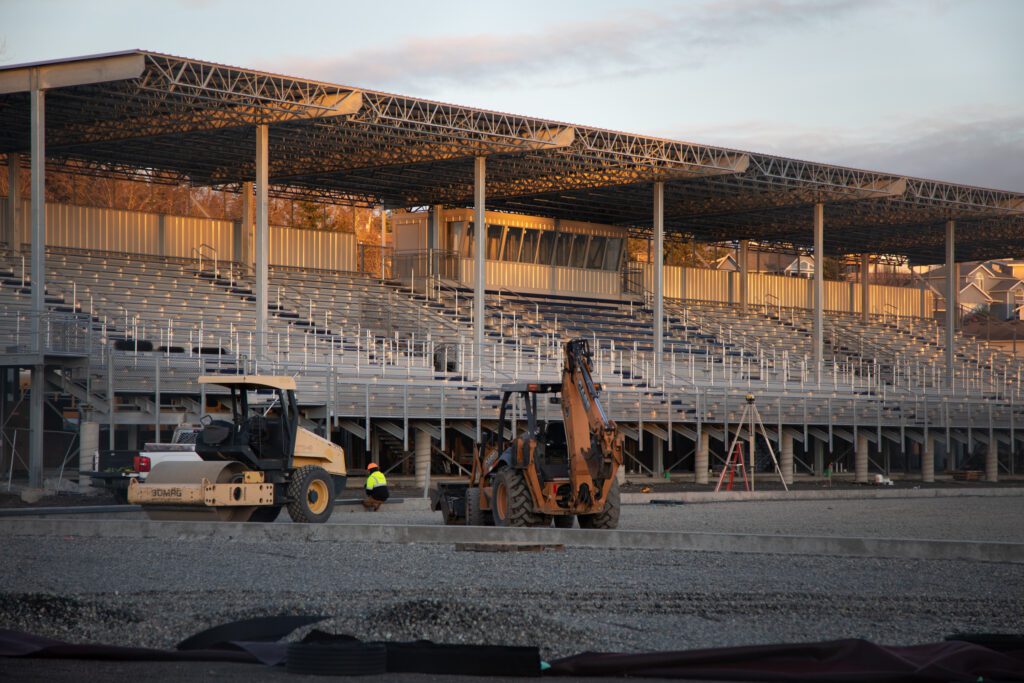 Construction vehicles and workers idle next to the football field and grandstands.