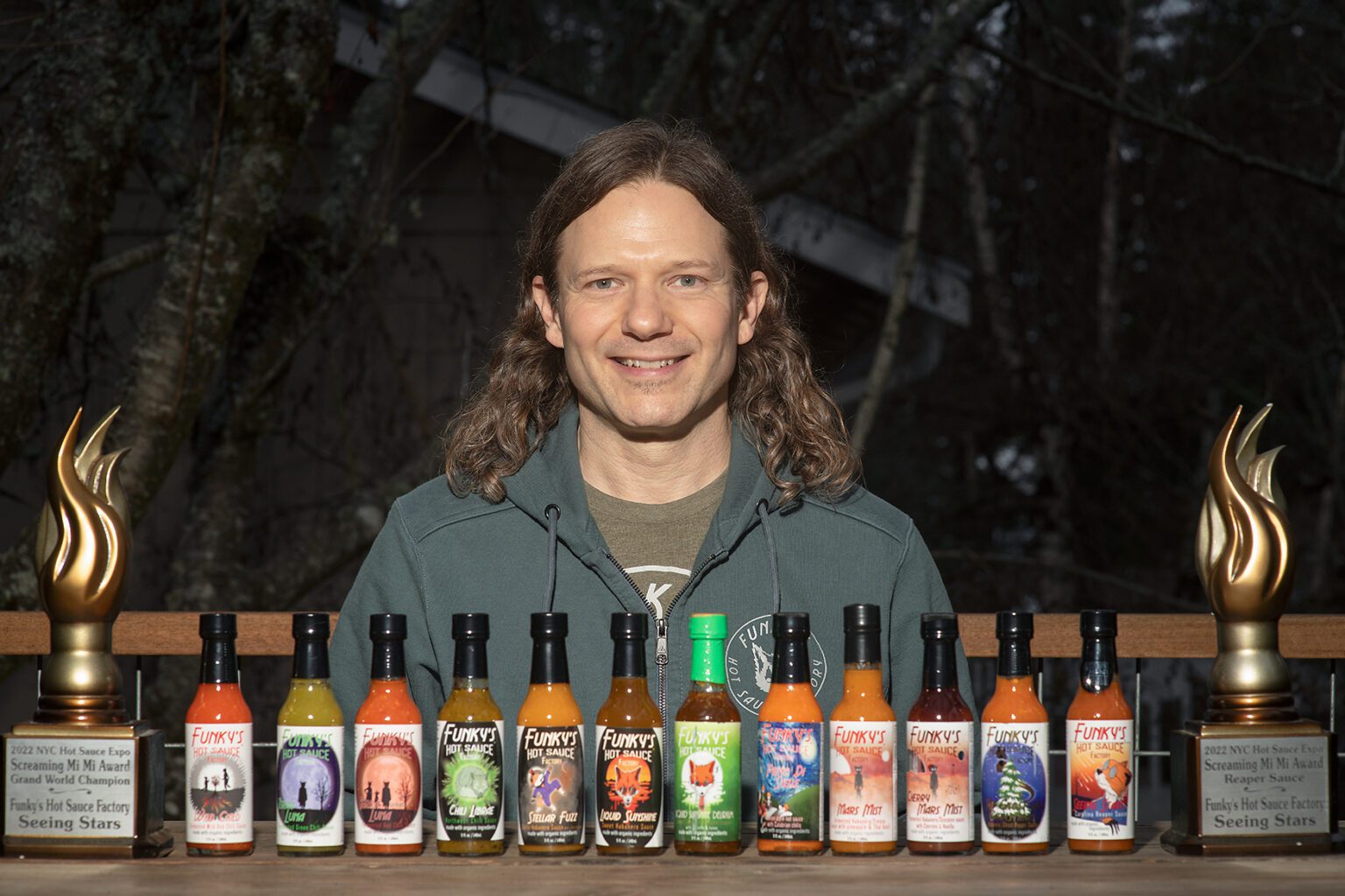 Matthew Mini smiling for the camera as he is posed behind the rows of hot sauce made by him.