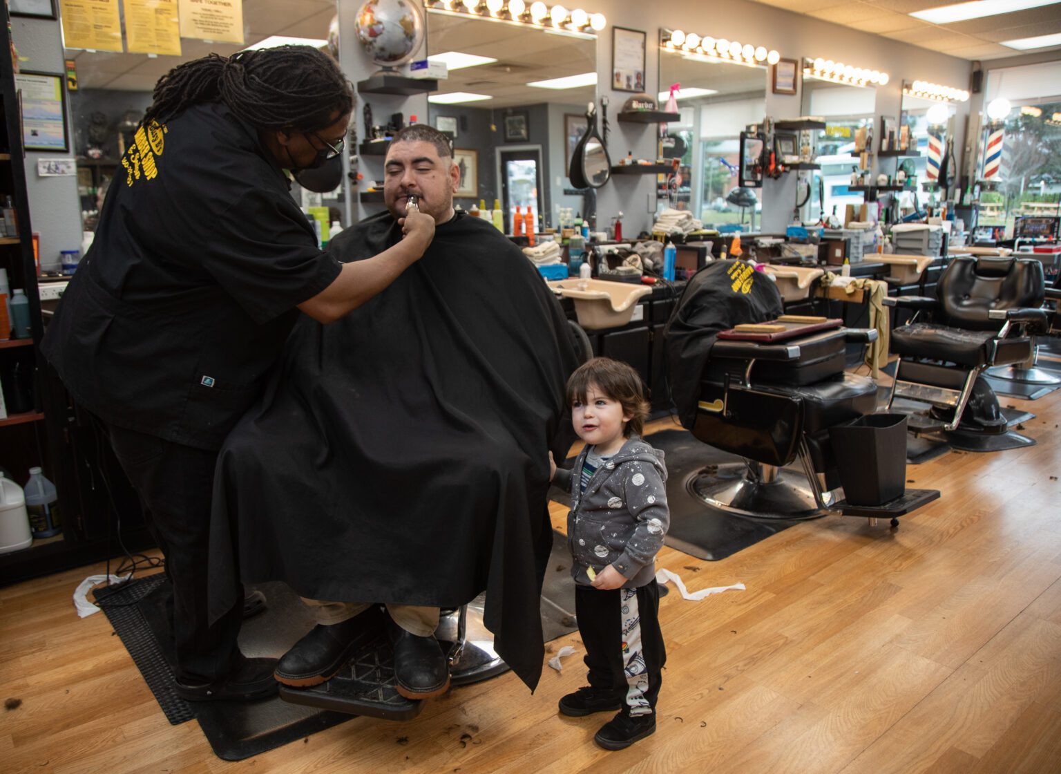 Bernard Franklin trims Fernando Martinez's mustache as his son, Niko, holding onto the barbershop's barber cape that's draped over his father.