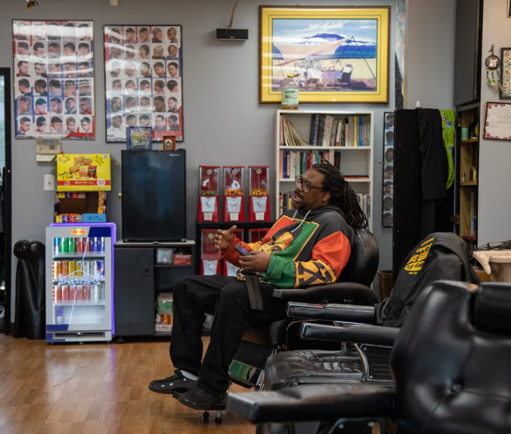 Bernard Franklin sits in his chair at his barbershop holding a remote and talking.
