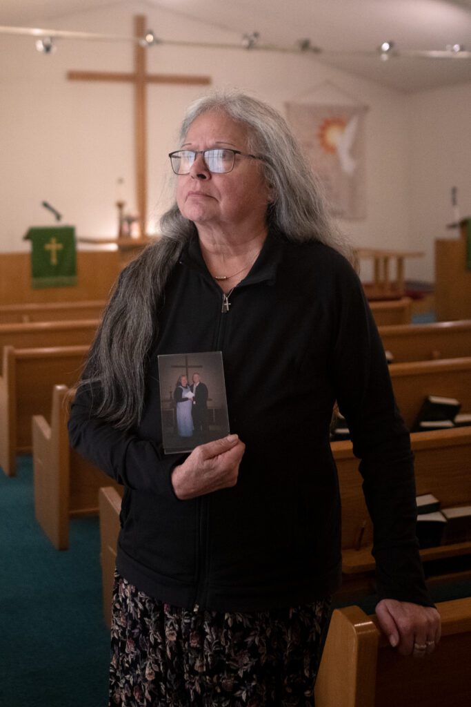 Nancy Perry holds a photo of her and her late husband, Rod Perry as she leans on the pews.
