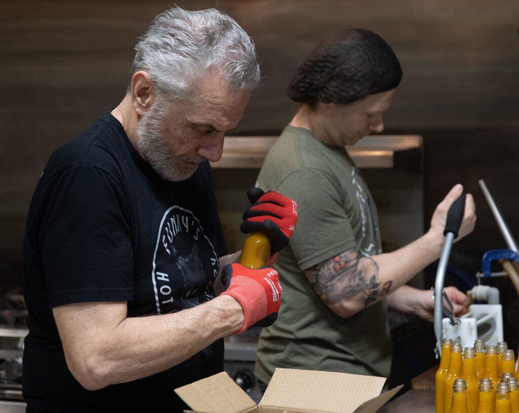 James Mini, Matthew's father, helps fill hot sauce bottles by hand as he looks down at the cardboard box to store.