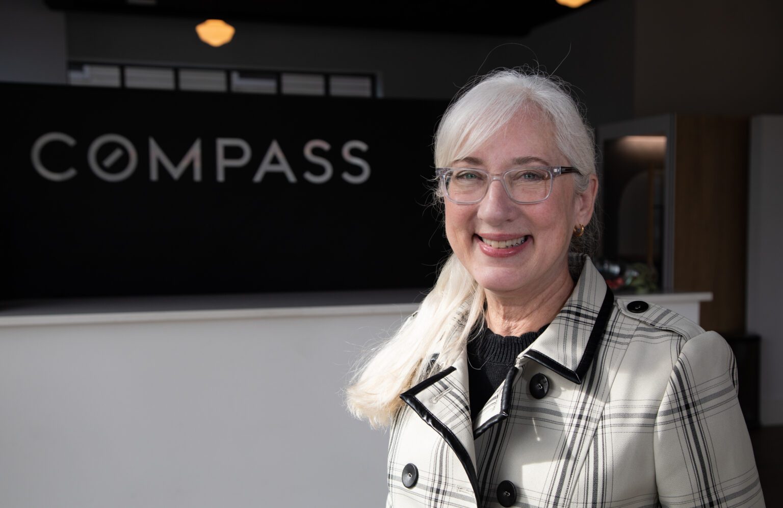A woman with glasses and a plaid jacket poses for a photo smiling in front of a wall that reads "Compass."