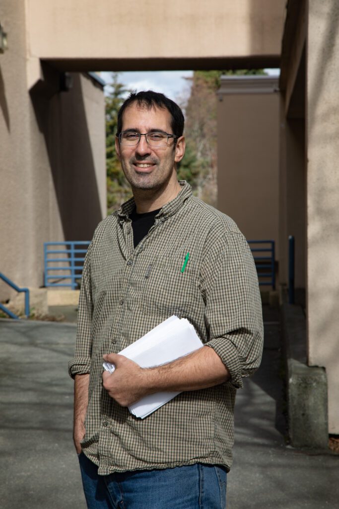A man wearing glasses and a plaid shirt poses for a photo holding a stack of papers.