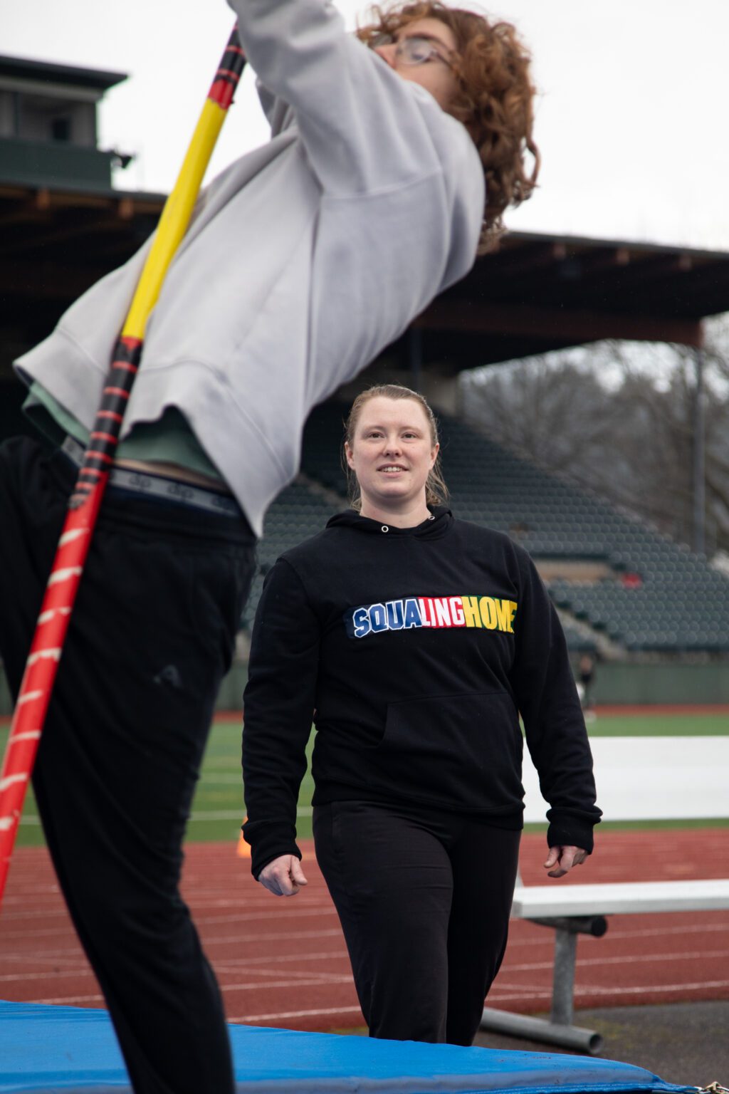 Dressed in her "SquaLingHome" sweatshirt, Coach Morgan Annable watches one of her students attempt to vault with a multi-colored pole.