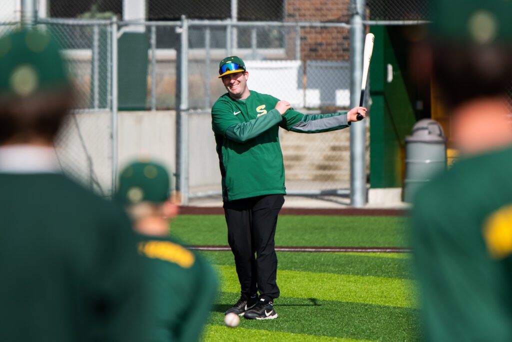 Dane Siegfried hits a groundball to infielders during practice with a smile on his face and dressed in the team's signature green uniforms.
