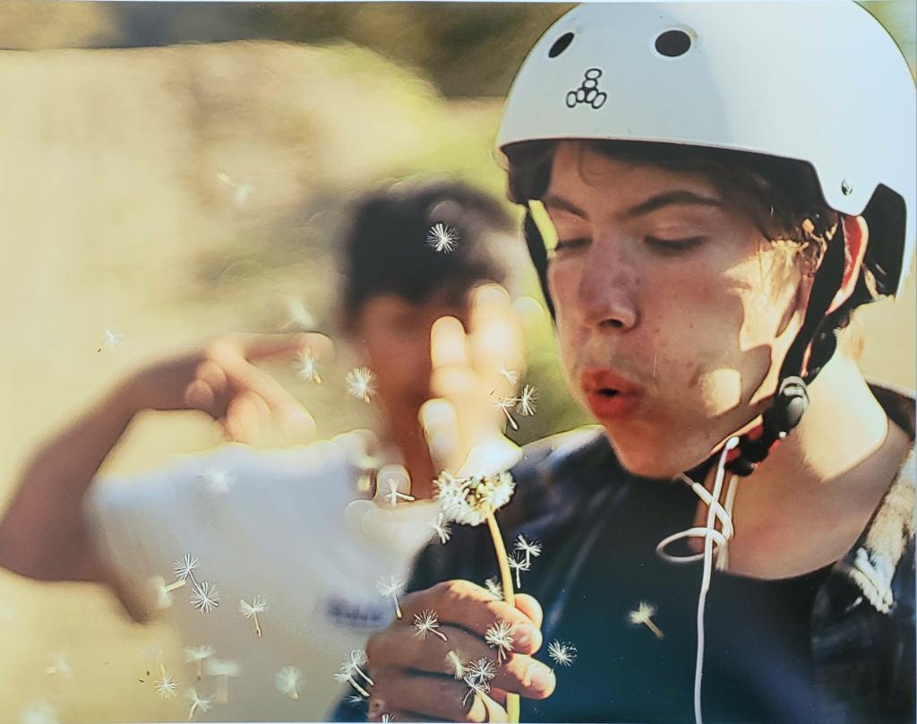 Aaren Coleman blows dandelion seeds while wearing a white bicycle helmet as another child is behind him, unfocused by the camera.