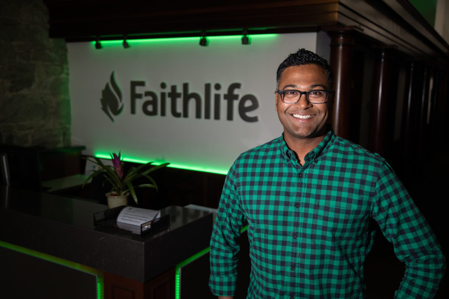A man wearing glasses and a green and black checkered shirt smiles for a photo in a green-lit room. The wall behind him reads "Faithlife."