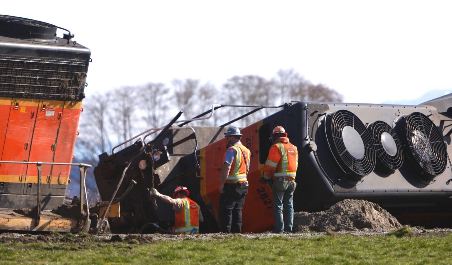 Crews examine an overturned locomotive wearing safety helmets and bright orange safety suits as they inspect deeper in the dirt.