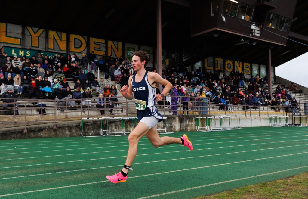 Lynden Christian's Andrew Luce running on the green track field as spectators from the bleachers watch.