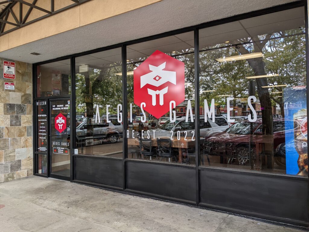 The storefront of Aegis Games showcasing their signature bright red logo on the glass windows.