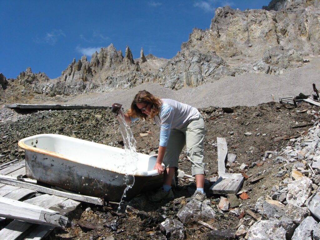 Anna Rankin pouring water out of a bathtub inbetween the rocky mountains surrounding the scrapyard.