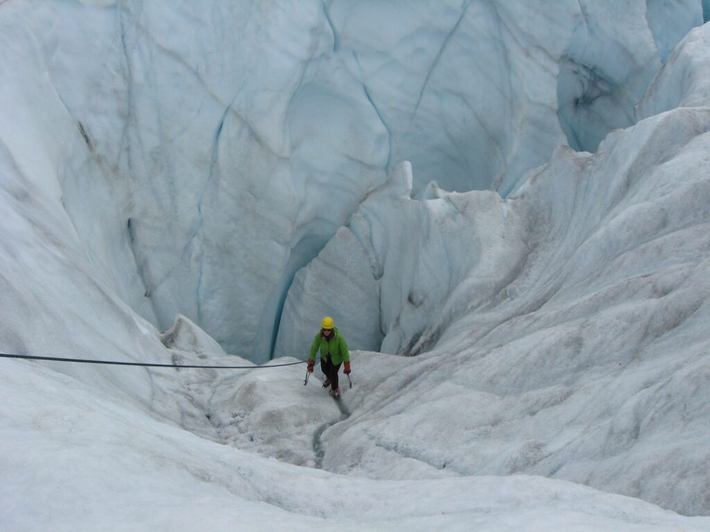 Anna Rankin ice climbs Kennicott Glacier with a rope tied to her to a higher elevated point while wearing a green vest and a bright yellow helmet.