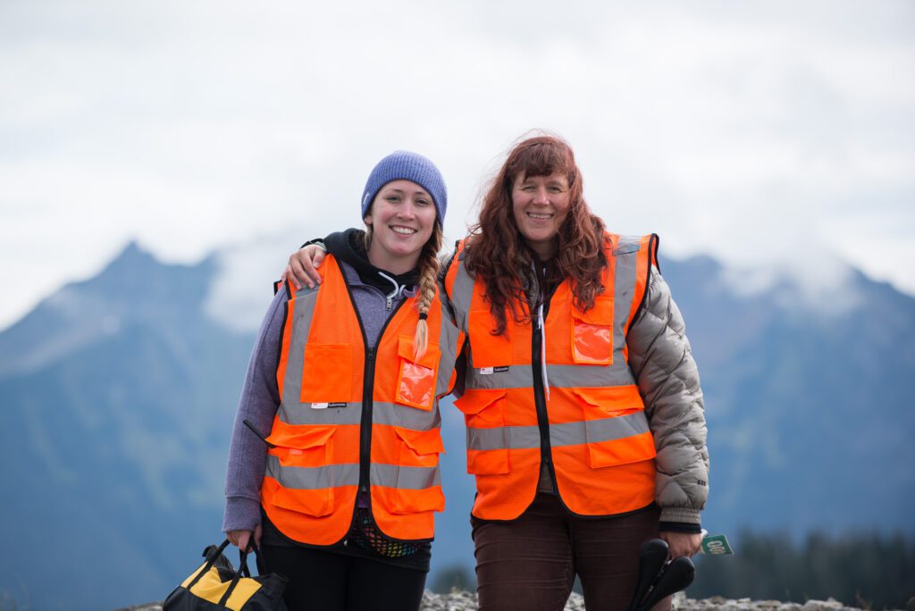 Former Assistant Race Director Ashlee Carstens, left, and Anna Rankin pose with an arm around their backs and the mountains in the background.
