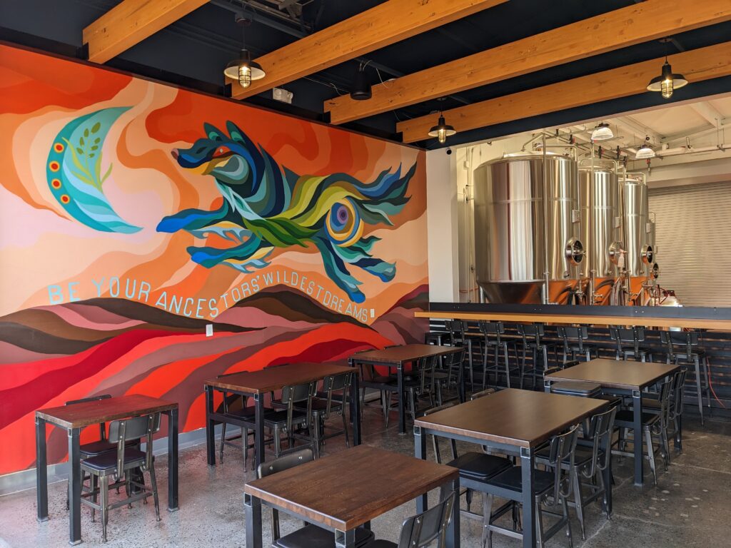 The interior of El Sueñito Brewing Company & Frelard Tamales with a colorful mural on the wall next to a bar top facing metal breweries.