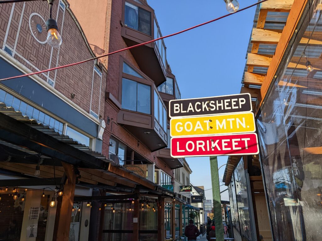 A black, yellow and red sign stacked on top of each other that reads "Blacksheep", "Goat MTN." and "Lorikeet" in between the buildings and a restaurant's outdoor seating space.
