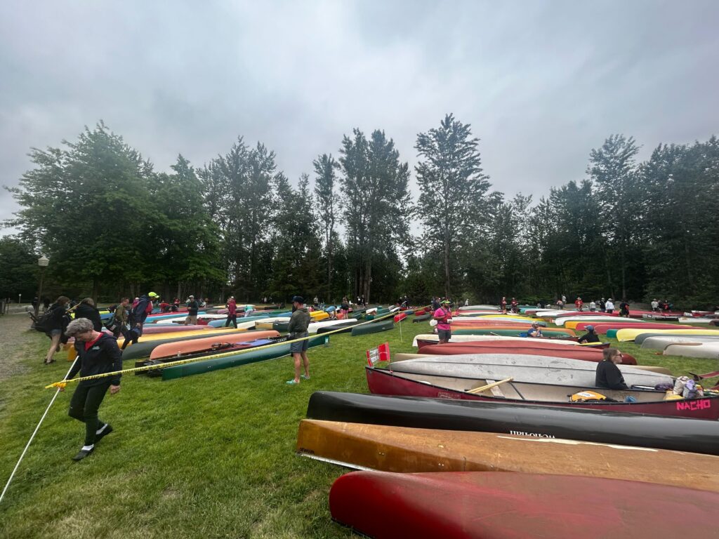 Canoeists and their canoes wait next to an array of colorful canoes.