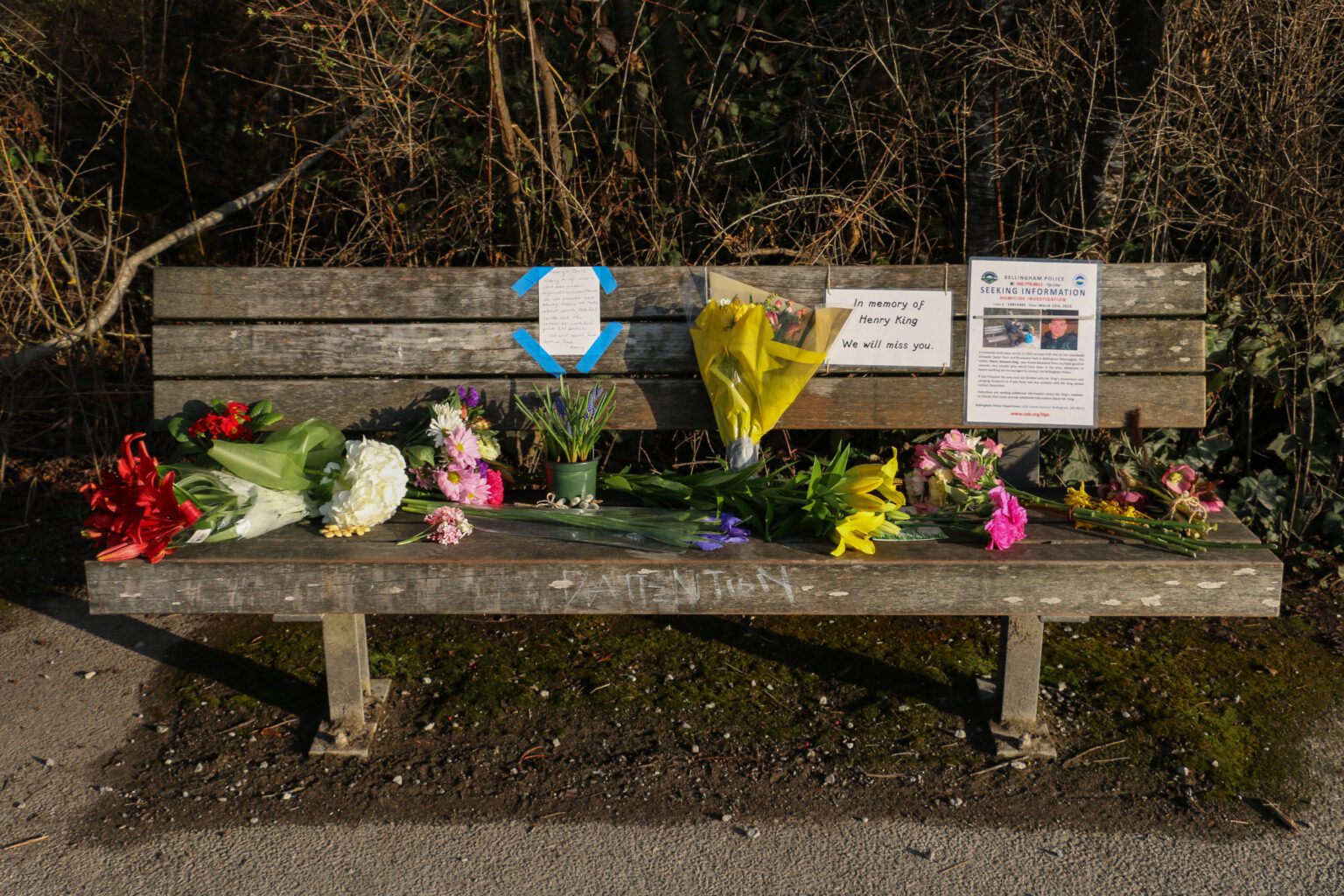 A wooden bench with many flowers left on top. There is an excerpt from the police for any information as well as a sign dedicating the bench in memory of Henry King.
