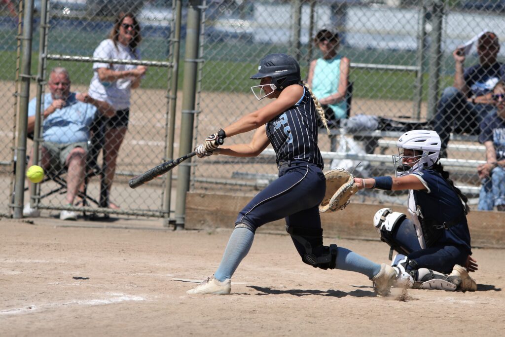 Lynden Christian's Kenadi Korthuis connects as she bats the ball while spectators watch from behind the chain-linked fence.