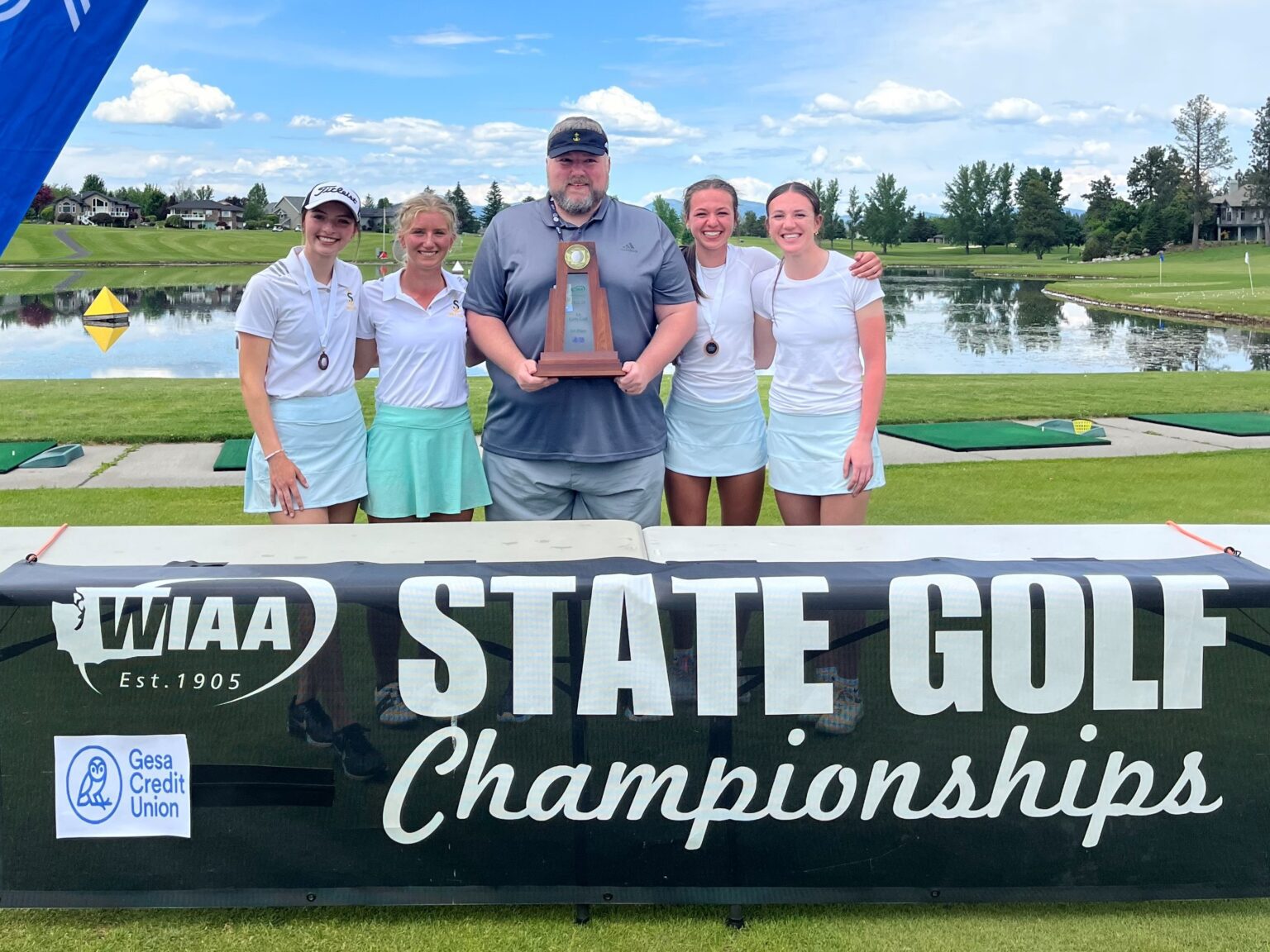 Sehome coach Jordan Chalfant posing for a photo with the trophy inbetween the Mariners girls golfers behind the state gold banner.